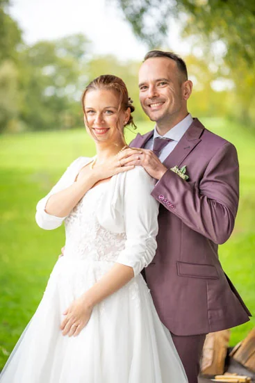 Braut und Bräutigam stehen lächelnd im Grünen, Bräutigam trägt einen bordeauxfarbenen Anzug